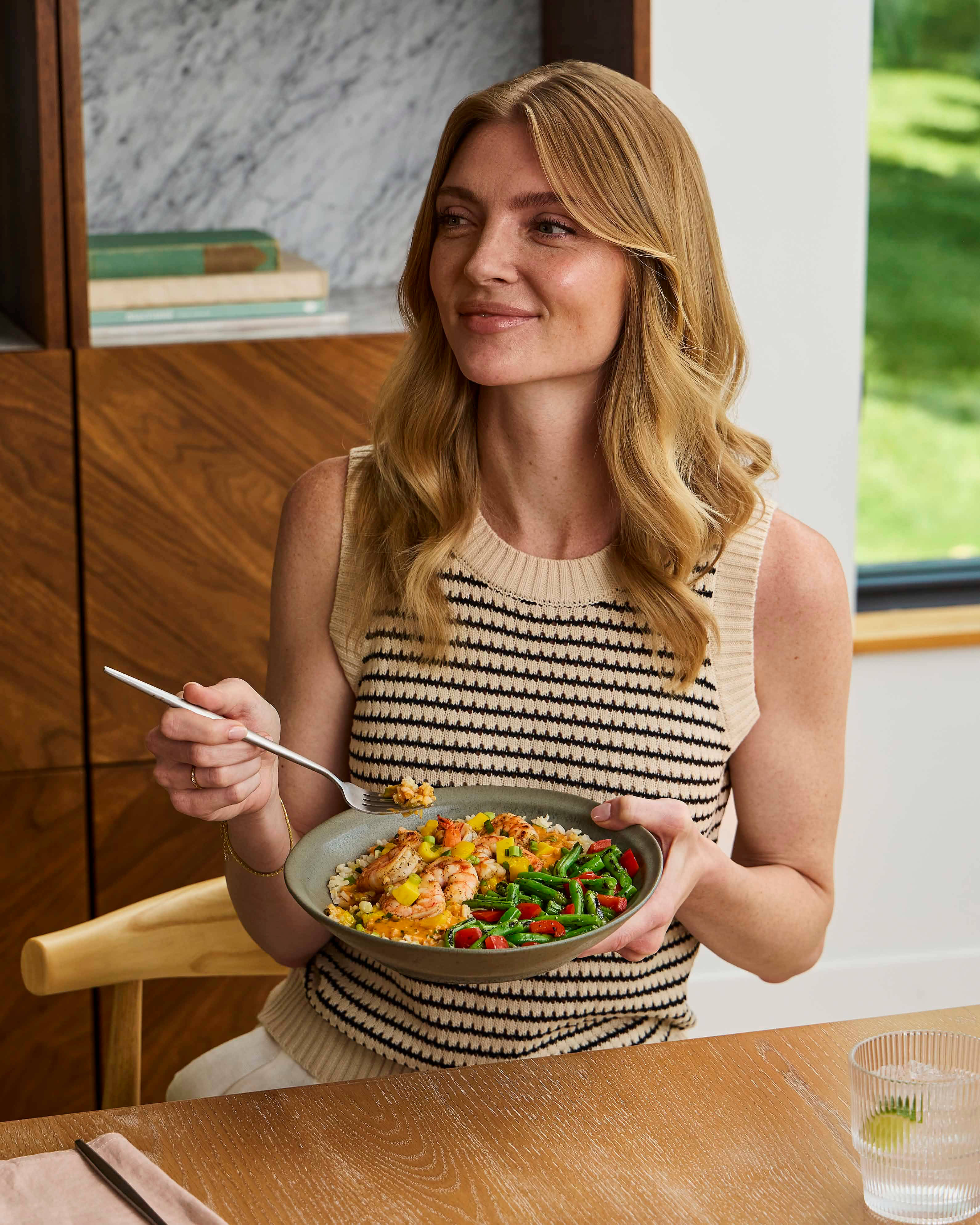 A Person smiling while holding a bowl of shrimp stir-fry with green beans, peppers, and rice.